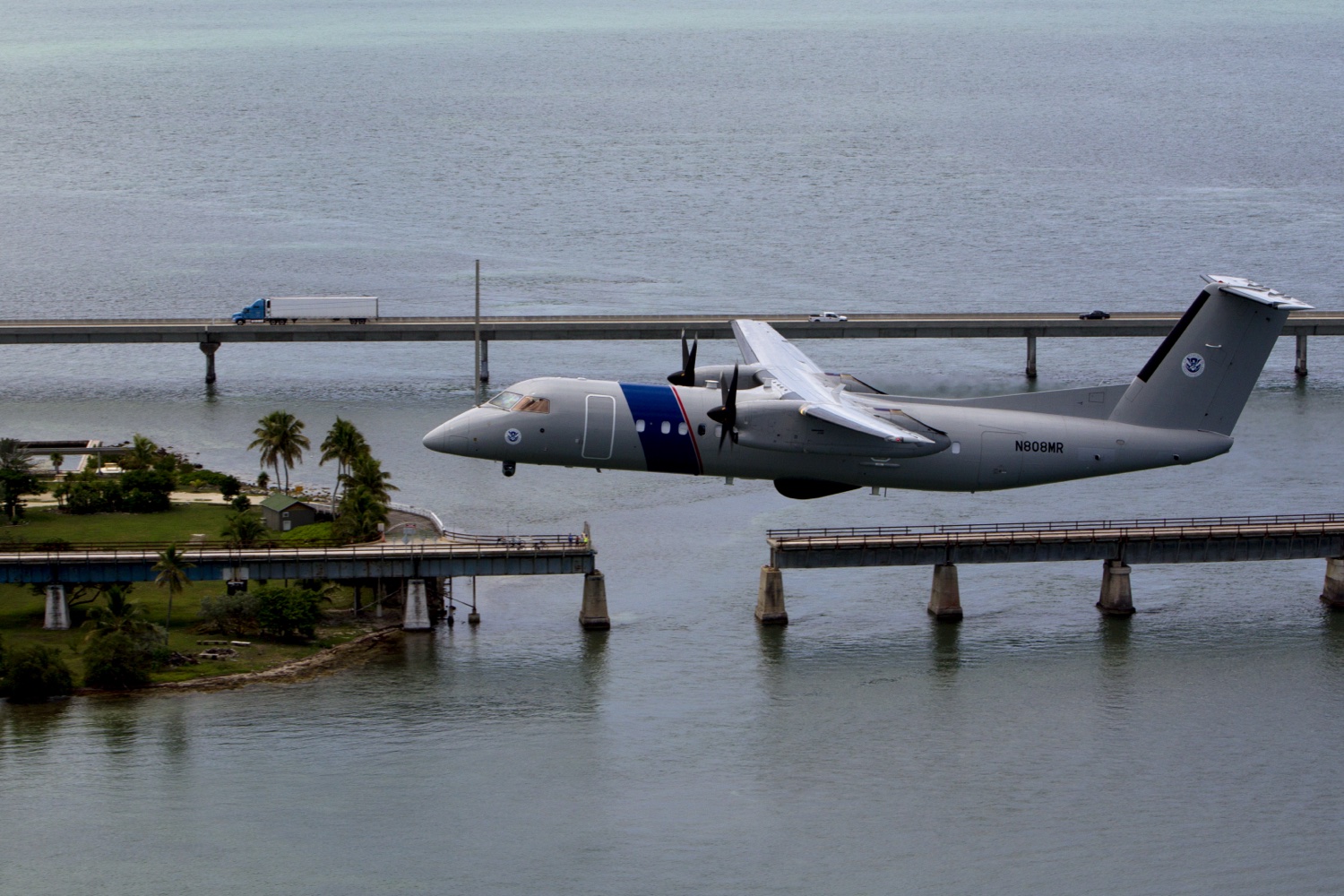 A U.S. Customs and Border Protection Office of Air and Marine Bombardier DHC-8 Dash 8 maritime patrol aircraft, tail N808MR, banking low over a Caribbean coastal bridge — the airframe class that carries the Wescam MX-15D FLIR turret used to record the Aguadilla incident.