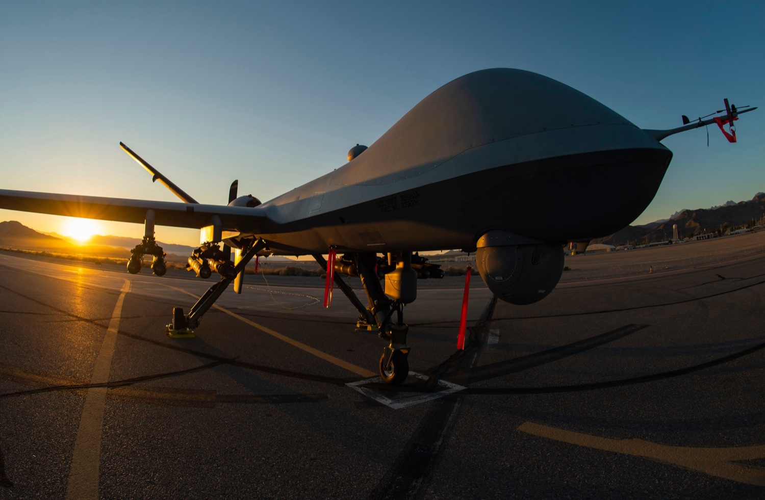 An MQ-9 Reaper sits on the flight line at Creech Air Force Base, Nevada, fully loaded with eight AGM-114 Hellfire missiles in the golden light of late afternoon — the airframe in profile, weapons pylons visible under both wings.