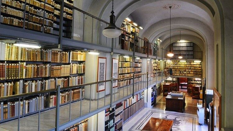 Interior of the Vatican Apostolic Library showing two-story shelving of leather-bound volumes, vaulted ceilings, hanging lamps, and a marble floor.